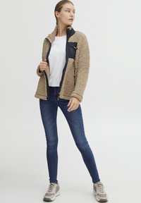 Veste en polaire beige avec une fermeture éclair noire marine et des poches, portée sur une chemise blanche. Jean skinny bleu foncé et baskets grises.