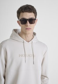 Sudadera beige con cordones, logo bordado "MORATO", combinada con gafas de sol negras. Material suave, diseño minimalista, ajuste casual.