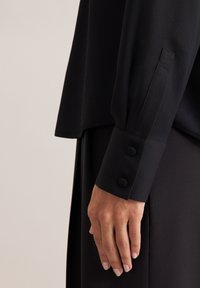 Blusa nera con una texture liscia e maniche lunghe, caratterizzata da un polsino con due bottoni rotondi. Primo piano di una mano appoggiata sul tessuto.