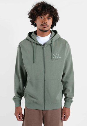 Sweat à capuche vert avec fermeture éclair, fabriqué en tissu doux. Comprend une poche avant, des cordons ajustables et un petit logo blanc sur la poitrine.