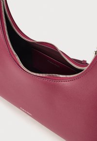 Calvin Klein táska mély bordó bőrből, sima textúrával, kerek formával és ezüst cipzáros részletekkel. Belsejében logó található.