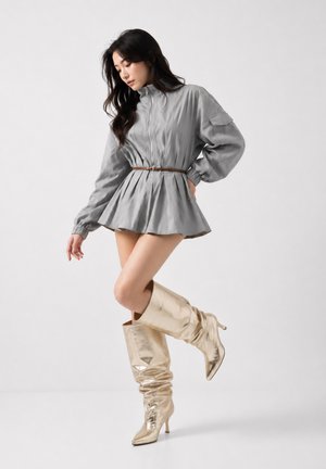 Femme portant une robe grise ceinturée à manches longues et des bottes dorées brillantes et amples montant jusqu'aux genoux, posant devant un fond clair uni.