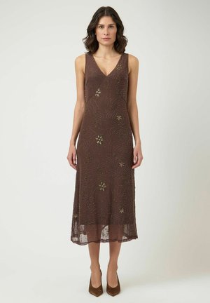 Nő ujjatlan barna midi ruhában, gyöngyös virágmintákkal és barna hegyes orrú magassarkú cipőben, sima háttér előtt áll.