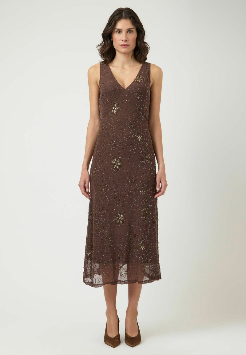 Femme portant une robe midi marron sans manches avec des motifs floraux perlés et des escarpins marron à bout pointu, debout devant un fond uni.