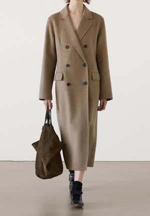 Femme portant un long manteau en laine camel croisé, tenant un sac en daim marron, avec des chaussettes et des chaussures noires, marchant sur un sol gris.