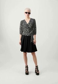 Schwarze Wickelbluse mit einem weißen geometrischen Muster, kombiniert mit einem schwarzen ausgestellten Rock. Das Modell trägt Plateausandalen und Sonnenbrille.
