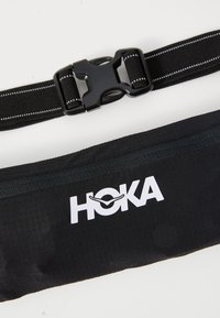 HOKA mustan juoksuvyön, joka on valmistettu pehmeästä kankaasta. siinä on vetoketjutasku, säädettävä hihna tukevalla muovisoljella ja valkoinen logo.