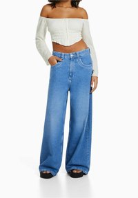 Donna che indossa un crop top bianco a spalle scoperte e jeans blu a vita alta e gamba larga, con le mani nelle tasche, in piedi su uno sfondo bianco.
