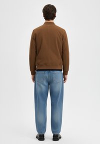 Giacca di cotone marrone con polsini a costine, abbinata a jeans denim azzurri chiari, larghi, con accenti sbiaditi e tasche posteriori.