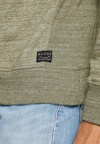 Felpa verde con trama a coste, caratterizzata da un'etichetta nera con "BLEND 1993" sul bordo, abbinata a jeans denim blu chiaro.
