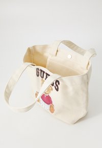 Sac en toile crème, comportant un graphique d'ours avec un haut rouge et une jupe blanche, avec "GUESS" imprimé en noir. Deux poignées pour le transport.