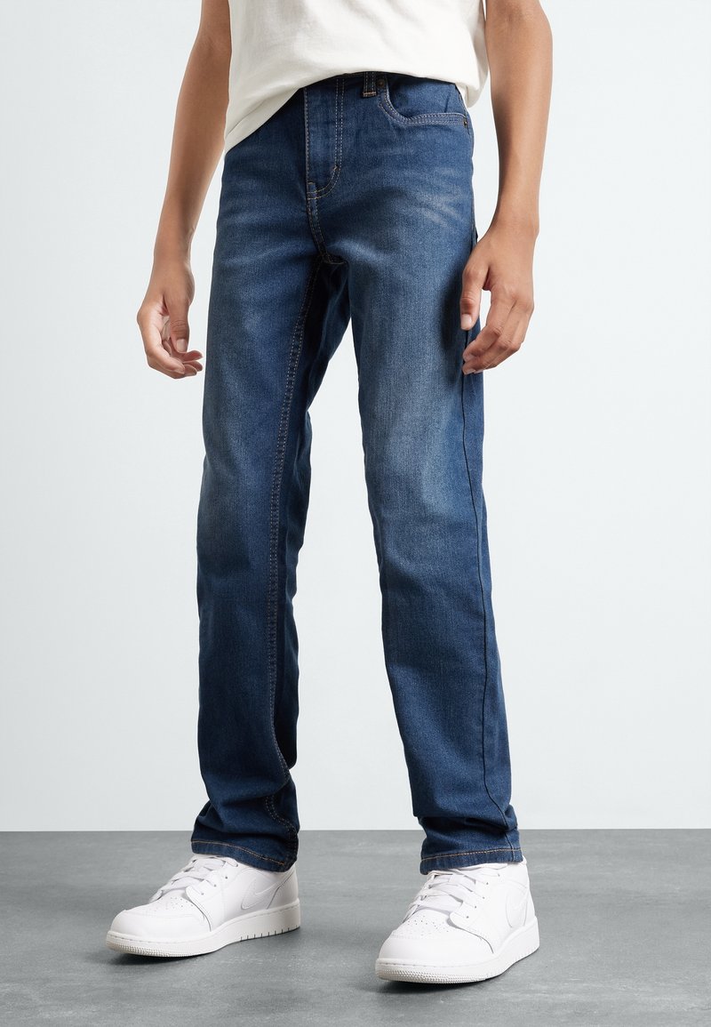 Jeans de mezclilla azul oscuro con corte recto, diseño de cinco bolsillos y acentos de costura, combinados con zapatillas blancas.