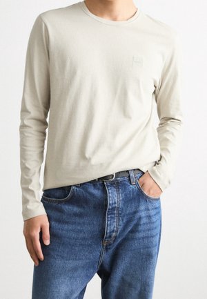 Man wearing a beige long-sleeve shirt tucked into blue denim jeans with a black belt, one hand in pocket, against a plain background.