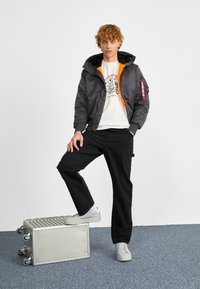 Black bomber jacket with orange lining, gray hoodie, graphic white t-shirt, black pants, and white sneakers, standing on a silver crate.