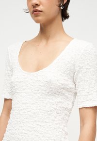 Femme portant un haut blanc texturé à manches courtes avec un col en U et des boucles d'oreilles argentées en forme d'anneau, sur un fond uni.