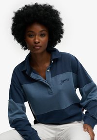 Kobieta o kręconych włosach, ubrana w niebieską koszulkę rugby Superdry i białe spodnie, siedząca na tle jednolitego białego tła.