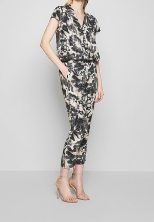 Woman wearing a black and white tie-dye jumpsuit with short sleeves and beige high-heeled sandals, standing with one hand in pocket.