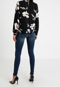 Blouse noire à motifs floraux avec des manches longues et un dos à découpe en clé, associée à un jean skinny bleu et des talons hauts noirs. Textures lisses, couleurs contrastées.
