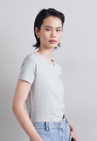 Tricou gri cu maneci scurte, fabricat din bumbac moale, cu guler rotund și un logo discret pe piept. Asortat cu blugi din denim de un albastru deschis.