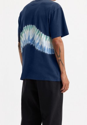 Osoba ubrana w granatowy T-shirt z zielonym, białym i niebieskim paskiem farbowanym na tkaninie z tyłu, w połączeniu z czarnymi spodniami, widoczne tatuaże na ramionach.