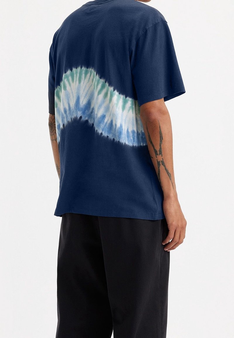Personne portant un t-shirt bleu marine avec une bande tie-dye verte, blanche et bleue à l'arrière, associée à un pantalon noir, tatouages visibles sur les bras.