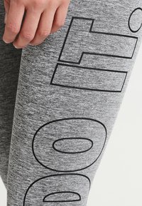 Szare legginsy sportowe z gładką fakturą i wyraźnym czarnym wzorem tekstowym na lewym udzie. Zbliżenie na dłoń trzymającą materiał.