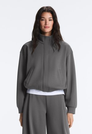 Vrouw draagt losse grijze jas met rits en hoge kraag en bijpassende wijde broekspijpen, witte blouse zichtbaar eronder.