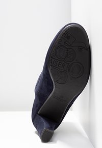 Chaussure à talon haut en daim bleu marine inclinée pour afficher une semelle noire avec des motifs de pièces embossés et le logo Peter Kaiser.