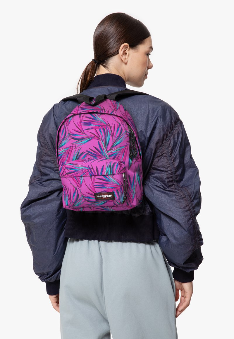 Zaino viola con un motivo di foglie tropicali, tasca frontale con zip e spallacci regolabili. Indossato sopra un giubbotto scuro e gonfio e pantaloni grigi.