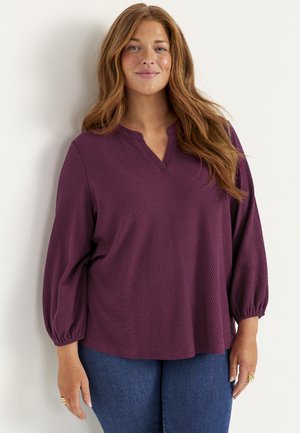 Vrouw met lang bruin haar, die een gestructureerde paarse blouse met driekwart mouwen en een blauwe spijkerbroek draagt, staat tegen een witte muur.