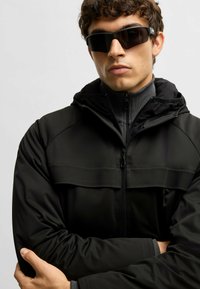 Svart zip-up jacka med en texturerad bröstpanel, parat med stilrena svarta solglasögon. Materialet verkar slätt och väderbeständigt.