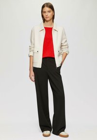Veste légère crème avec poches plaquées, associée à un haut rouge, un pantalon large noir et des chaussures beiges. Tissu lisse, coupe décontractée.