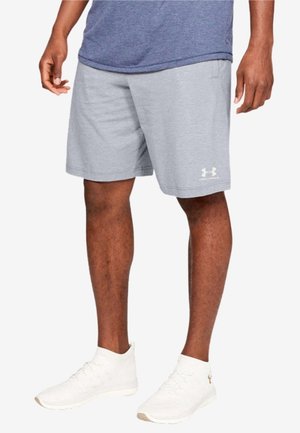 Man draagt lichtgrijze Under Armour short, blauw T-shirt en witte sportschoenen, staand tegen een witte achtergrond.