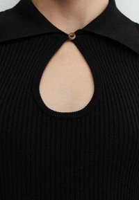 Top noir à côtes avec une découpe en forme de clef au niveau de la poitrine, accentué d'un bouton rond marron et d'un col replié.