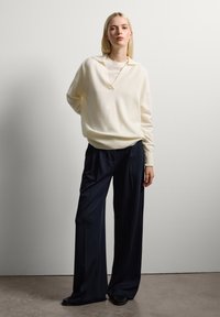 Cremfarbener, weicher Pullover mit Kragen, über einem weißen Oberteil getragen. Kombiniert mit navyblauen, weit geschnittenen Hosen und schwarzen Schuhen.