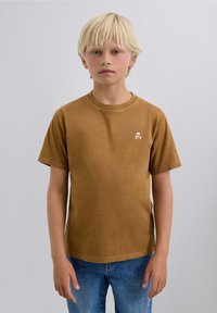 Camiseta de algodón marrón con un clásico escote redondo, mangas cortas y un logotipo de calavera y huesos cruzados en blanco en el lado izquierdo del pecho.