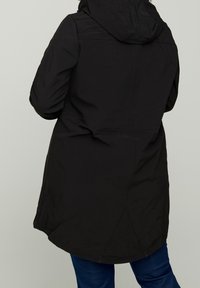 Parka noire imperméable avec capuche, présentant une taille ajustée, une coupe mi-longue et une texture lisse. Les détails des coutures à l'arrière sont visibles.