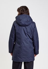 Femme vue de dos portant un manteau d'hiver à capuche bleu foncé et un pantalon sombre sur un fond uni.
