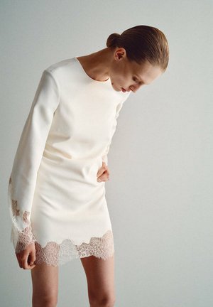 Mujer que lleva un vestido blanco de manga larga con ribete de encaje, mirando hacia abajo y sujetando su cintura sobre un fondo liso y claro.