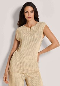 Vrouw met lang, donker haar, gekleed in een mouwloos beige textuurtop en bijpassende broek, poseert met één hand op de heup tegen een lichte achtergrond.