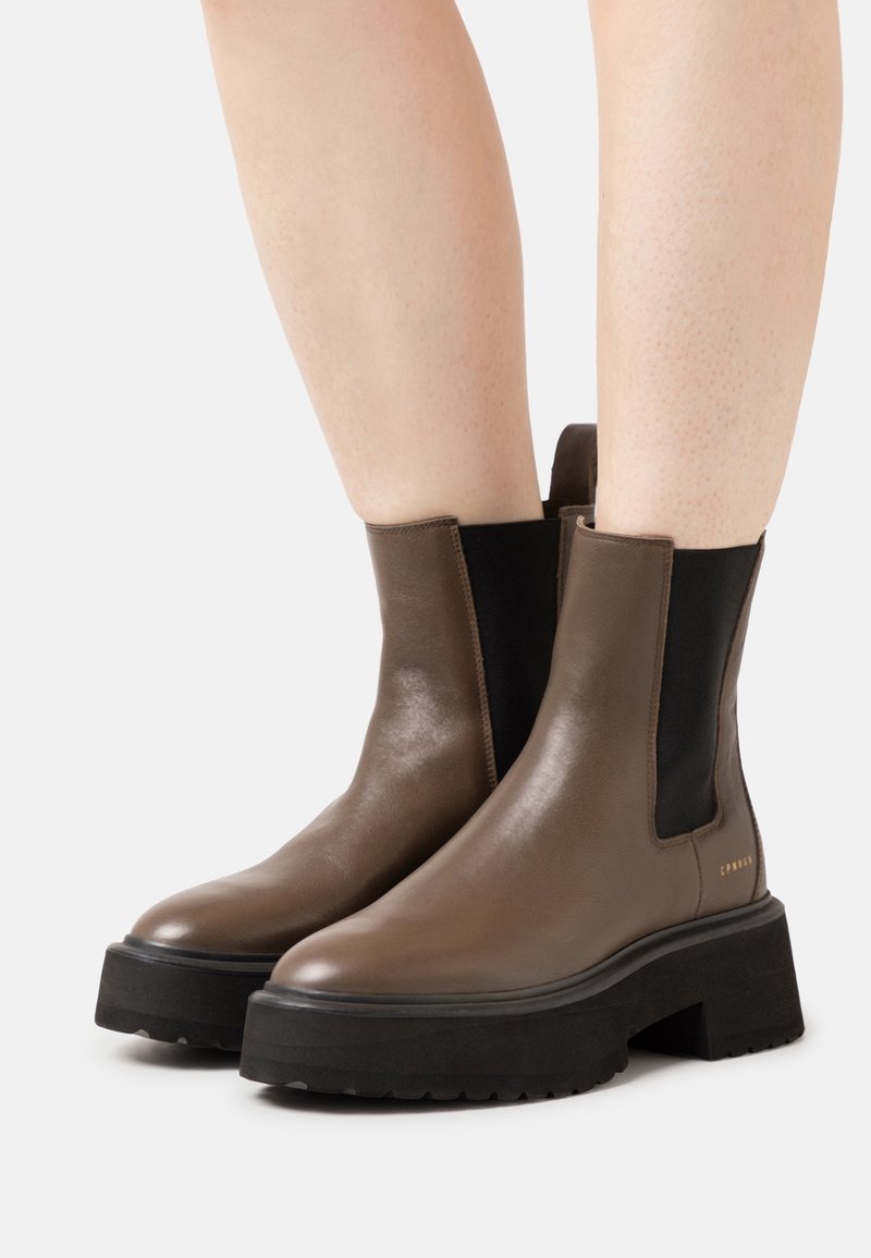 Bottines en cuir marron avec semelles épaisses noires et panneaux latéraux élastiques portées sur des jambes nues sur un fond uni.