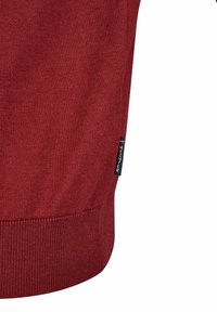 Červený pletený svetr s manžetami a spodním lemem se žebrováním. Výrazná značka na boční straně s horizontálním pruhovaným akcentem. Hladká textura.