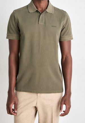 Mann trägt olivgrünes kurzärmeliges Poloshirt mit "BOSS"-Logo und beige Hose vor schlichten hellem Hintergrund.