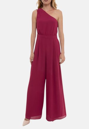 Donna che indossa una tuta senza maniche, a una spalla, di colore rosa intenso con pantaloni ampi e tacchi chiari a punta su uno sfondo bianco uniforme.