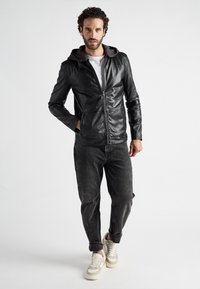 Svart läderjacka med dragkedja, med en grå huva och två framfickor, bärs tillsammans med blekta svarta jeans och ljusa sneakers.