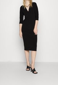 Svart figurnära klänning med V-ringning och trekvartsärmar, tillverkad av ett mjukt tyg, kombinerad med svarta sandaler med öppen tå.