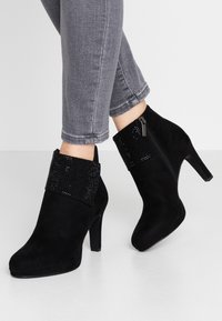 Mustat mokkasaappaat, joissa on korkea korko ja kimalteleva rannetyyli, yhdistettyinä harmaisiin kireisiin farkkuihin, seisoo vaalealla lattialla.