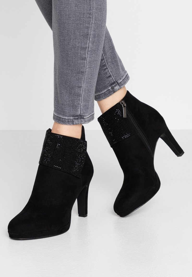 Mustat mokkasaappaat, joissa on korkea korko ja kimalteleva rannetyyli, yhdistettyinä harmaisiin kireisiin farkkuihin, seisoo vaalealla lattialla.