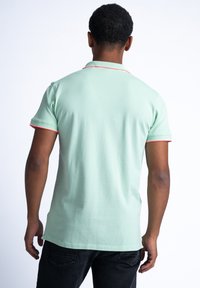 Jasnozielona koszulka polo o miękkiej teksturze, z krótkimi rękawami i czerwonymi paskami na kołnierzyku oraz końcach rękawów. Minimalistyczny design.