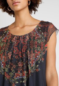 Blomstret kjole med korte, gennemsigtige ærmer. Har mørkt stof med røde, grønne og blå blomster samt et indsamlet halsudskæringsdesign.